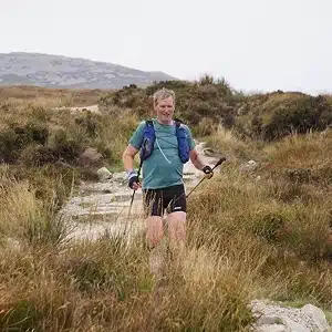 A person wearing a blue top and shorts hikes with trekking poles on a rocky trail surrounded by tall grass and hills under a cloudy sky.