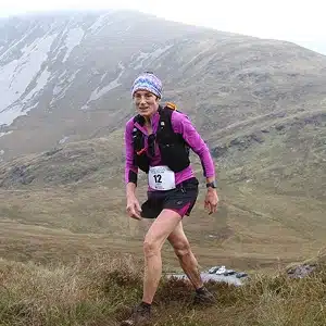A runner wearing a purple shirt, black shorts, and a race bib number 12 climbs a grassy, hilly landscape with misty mountains in the background.