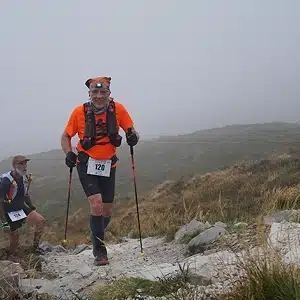 Two runners with race bibs and trekking poles climb a rocky mountain trail on a foggy day. The lead runner wears an orange shirt and black shorts, while the other follows closely behind. Grassy hillside and mist in the background.