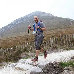 A man wearing athletic gear and using trekking poles hikes on a rocky trail with a grassy hill and a wooden fence in the background. He is smiling at the camera.
