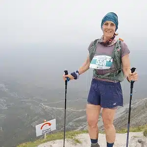 A woman with hiking poles stands on a mountain trail, wearing a race bib numbered 558, shorts, and a headscarf. There is a sign on the ground nearby, and the background shows a misty, expansive landscape.