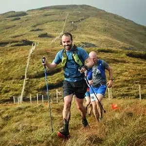 Two men are trail running up a grassy, muddy hillside using trekking poles. The runner in front wears a green vest and black shorts, while the second wears blue. More runners follow in the distance under a cloudy sky.