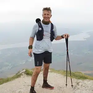 A man wearing a hydration vest, t-shirt, and shorts stands on a mountaintop holding trekking poles, with muddy legs and a scenic landscape of hills, water, and sky in the background.