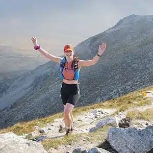 A woman in athletic wear and a cap is hiking on a rocky mountain trail with her arms raised. The background shows a scenic, grassy landscape and mountain slopes under a hazy sky.