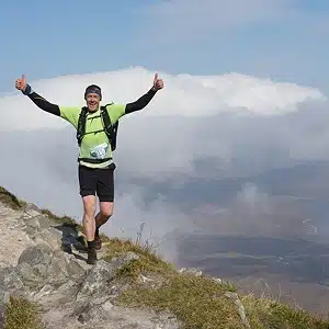 A man in hiking gear and a race bib gives two thumbs up while running along a rocky mountain trail, with clouds and a scenic landscape in the background.