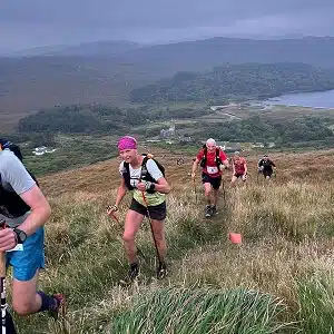 A group of trail runners with race bibs climb a grassy hillside under a cloudy sky, with a lake and forested hills visible in the background. One runner in front wears a pink headband and carries trekking poles.